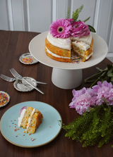 The Tiny Finch sweet cream confetti cake on a white pedestal, topped with pink flowers and a slice on a blue plate.