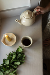 The Tiny Finch handmade cream stoneware teapot with matching cup, candle holder, and fresh mint on a grey surface.
