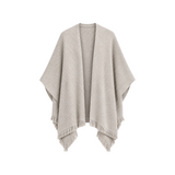 The Tiny Finch Cape Sand is a beige knitted open-front poncho featuring a textured ribbed pattern and fringe hem.