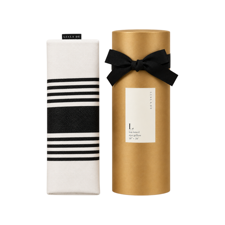 The Tiny Finch lavender eye pillow in white with black stripes, shown next to its gold gift tube with a black bow.