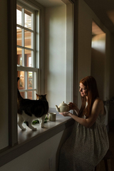 The Tiny Finch handmade stoneware teapot held by a woman sitting by a window with her cat in a cozy home setting.