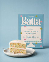 The Tiny Finch Batta Sweet Cream Confetti cake mix box next to a slice of frosted confetti cake on a plate.