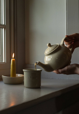 The Tiny Finch stoneware teapot pouring tea into a matching mug beside a lit candle on a white background