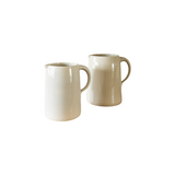 Two The Tiny Finch handmade stoneware pitchers in cream on a wooden shelf in a farmhouse kitchen setting.