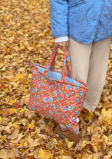 The Tiny Finch Persimmon quilted tote bag with floral print in red, blue, and green held against autumn leaves.