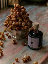 The Tiny Finch barrel-aged balsamic vinegar in a glass bottle next to a silver bowl of glazed popcorn on marble