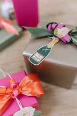 The Tiny Finch champagne bottle gift tag with gold foil accents and ribbon, tied to a metallic gold gift box.