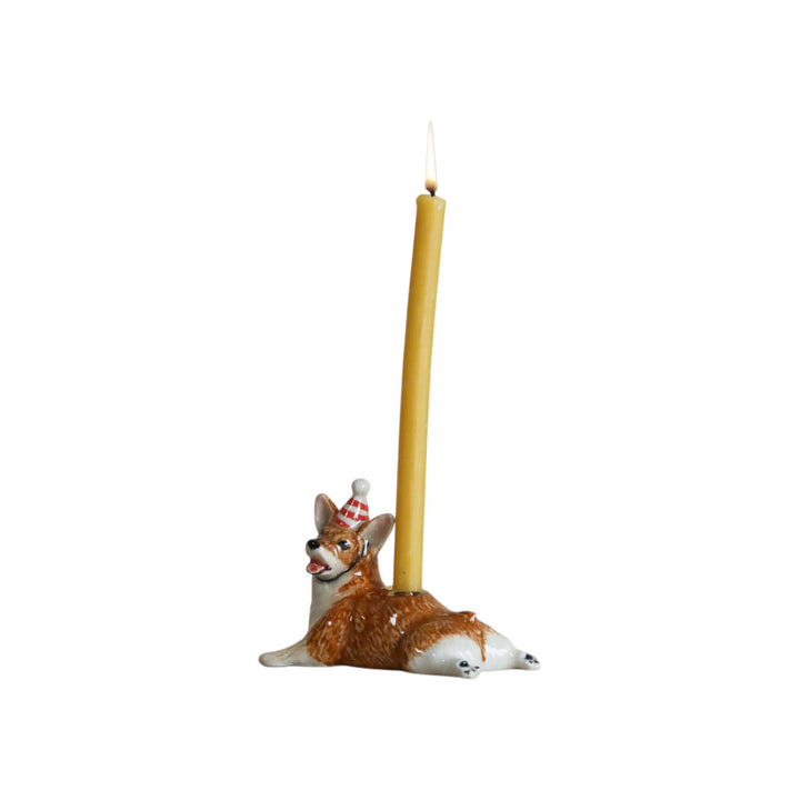 The Tiny Finch porcelain Corgi cake topper wearing a red and white striped party hat with a lit yellow candle.