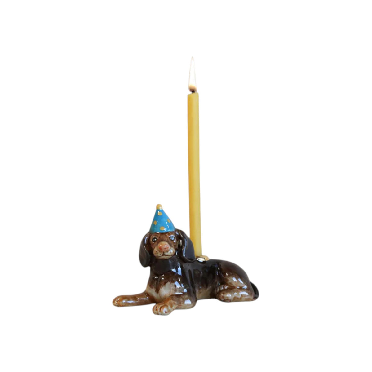 The Tiny Finch hand-painted porcelain dachshund cake topper wearing a blue party hat with a lit yellow candle.