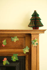 The Tiny Finch hand-printed holly and ivy paper garland draped across a wooden fireplace mantel with a paper tree.