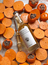 The Tiny Finch raw persimmon vinegar bottle surrounded by sliced Fuyu and Hachiya persimmons on a marble surface.