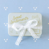 The Tiny Finch Sweet Sentiments keepsake tin with gold lettering and a white lace ribbon on a blue patterned background